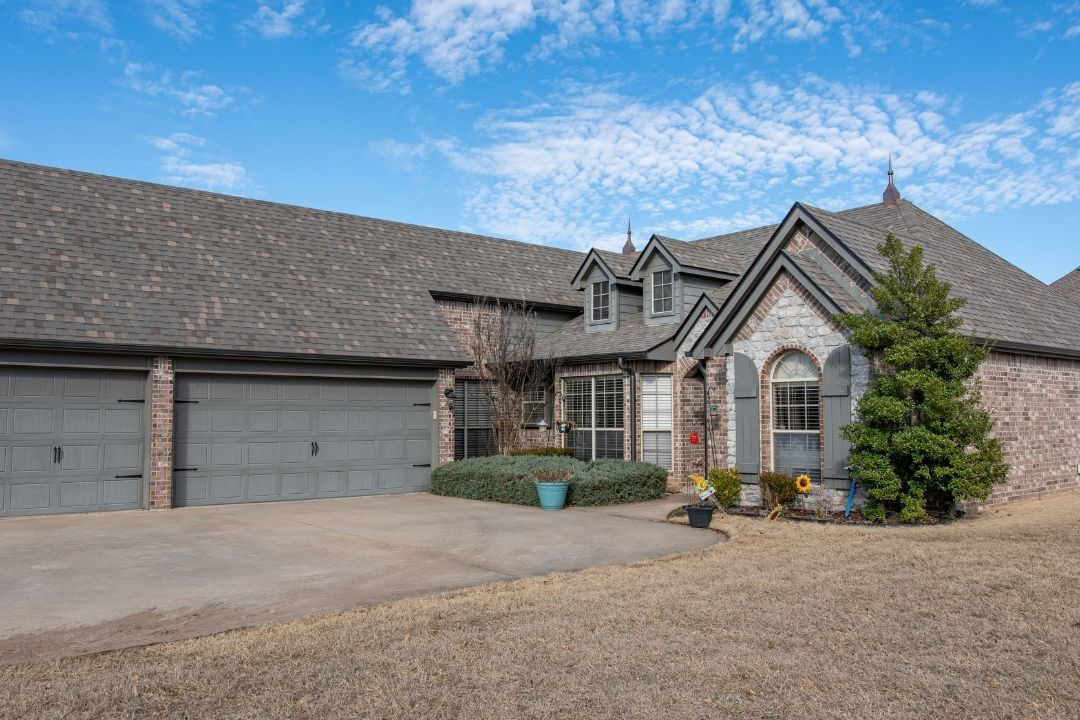 Completed dark shingle roof on upscale Tulsa brick and stone home — triple garage, dormers, decorative finials, gray shutters, blue Oklahoma sky