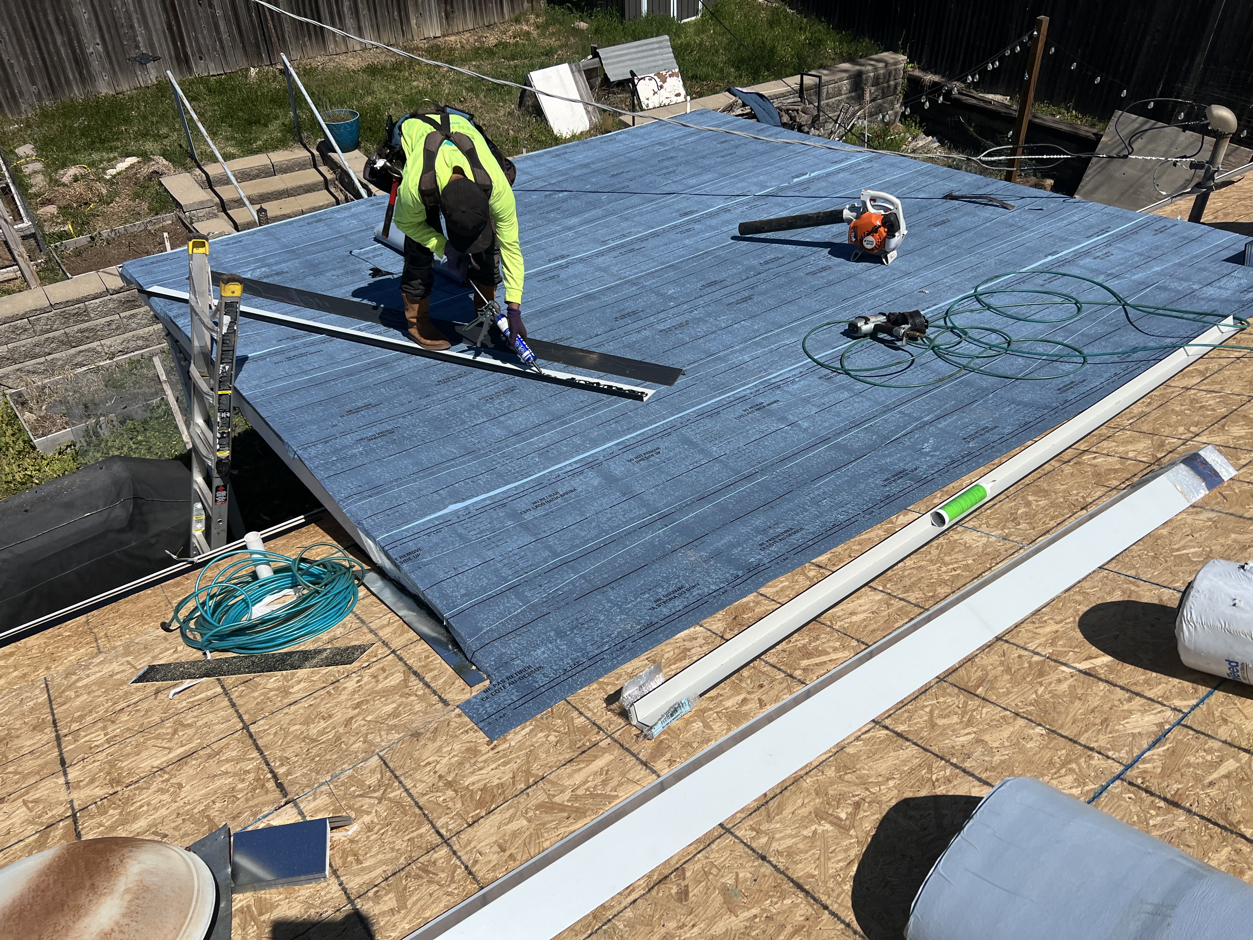 Crew member in GCR lime green laying underlayment on fresh OSB — half deck covered in blue synthetic, Stihl blower, air hose, drip edge being set