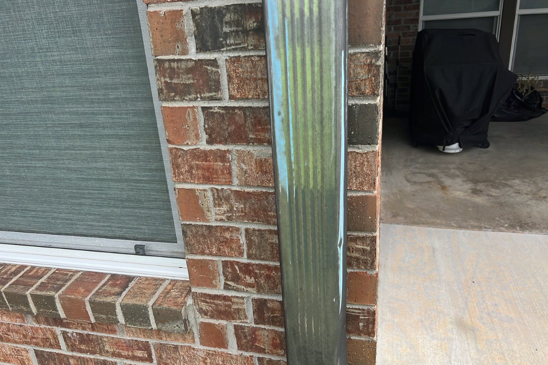 Hail dents on downspout with blue/green chalk marks — dings running down metal, brick home, patio visible