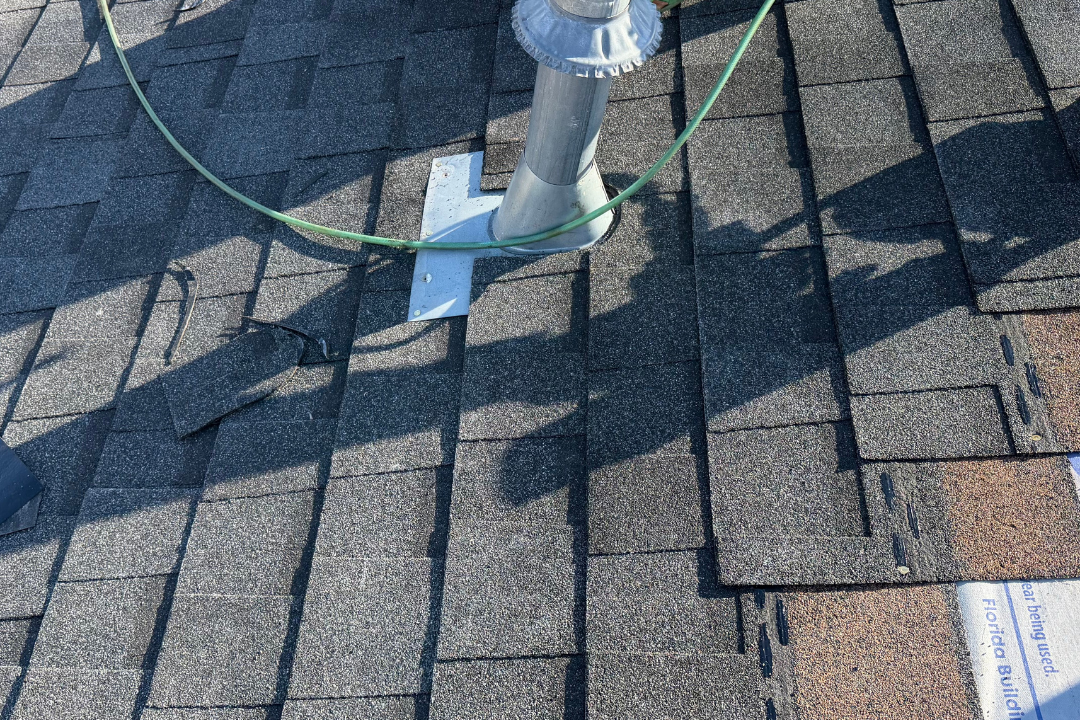 Pipe jack being installed on dark shingles — metal base plate, galvanized pipe boot, crew boots and green gloves, air hose, underlayment backing visible
