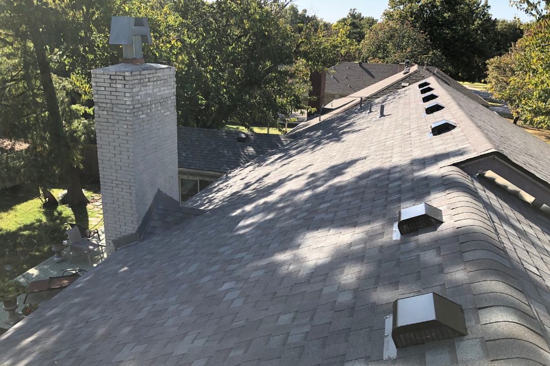 Aerial view large complex gray shingle roof — white brick chimney with copper cap, box vents, skylights, hip and valley transitions, mature Tulsa trees