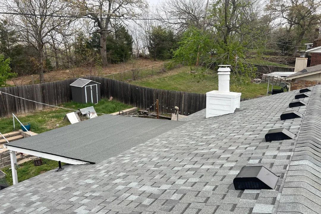 Aerial gray shingle roof — multiple box vents, white painted chimney chase, flat roof addition section, Tulsa spring trees budding