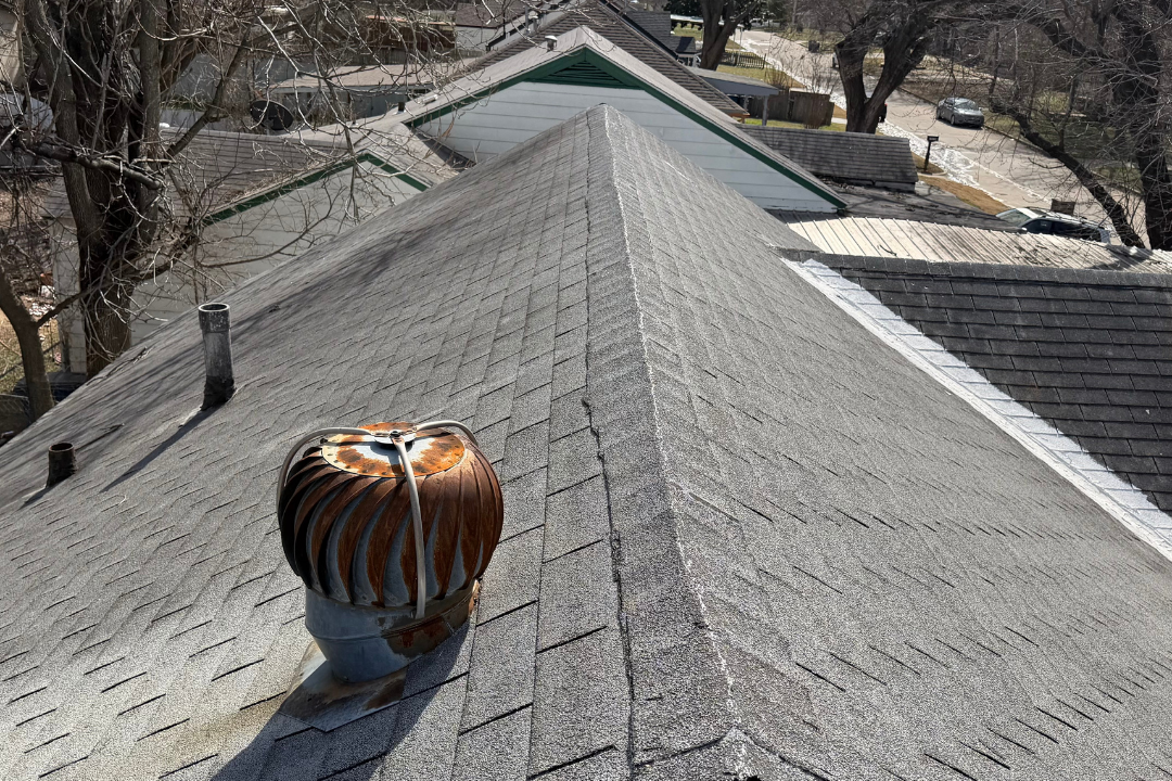 Rusted turbine vent on badly deteriorated roof — stripped shingles up the ridge, bare ridge cap, peeling eave shingles bottom right, Tulsa neighborhood