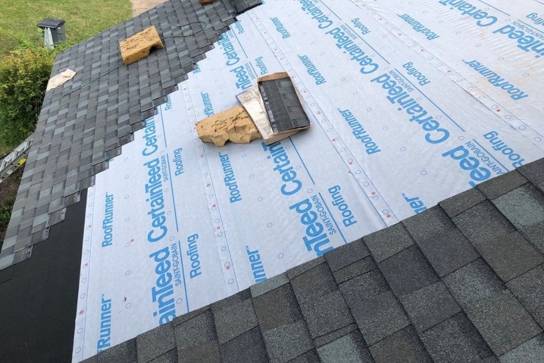CertainTeed RoofRunner underlayment mid-install — dark shingles going down both sides, shingle bundles staged on roof, crew boots visible top, van in driveway