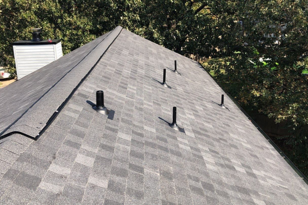 Pristine gray shingle roof from above — four black pipe jacks, continuous ridge cap full length, tight drip edge, mature Tulsa oak trees, detached garage