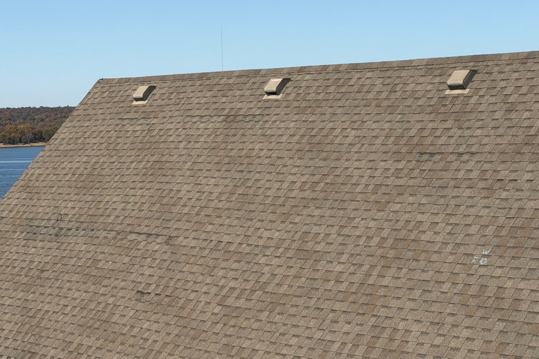 Aged roof with multiple wind damage spots across field — dark patches where shingles displaced, three box vents, chalk mark, Oklahoma lake backdrop