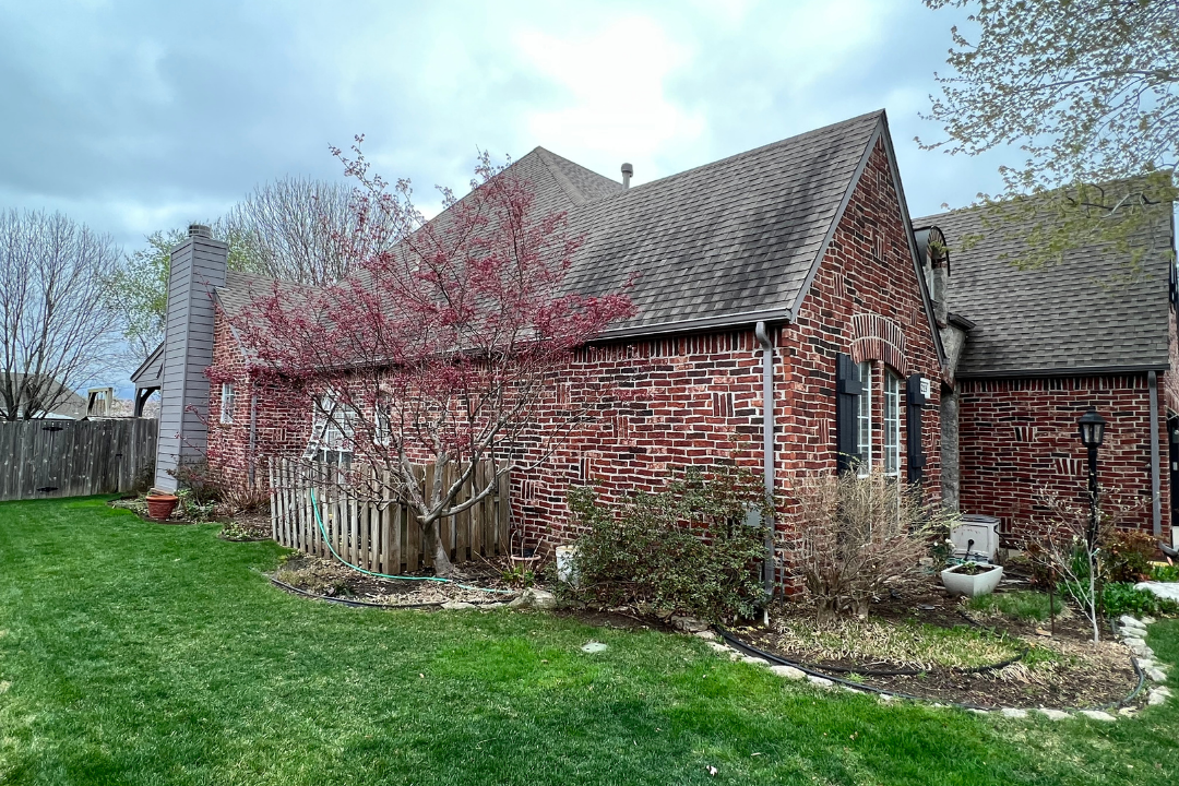 Algae streaks running down roofline of Tulsa brick home — spring blooming tree, green grass, dramatic cloudy sky