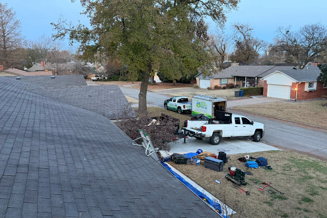 GCR crew member on roof — truck and trailer staged in driveway, tools laid out, Tulsa neighborhood, dusk light