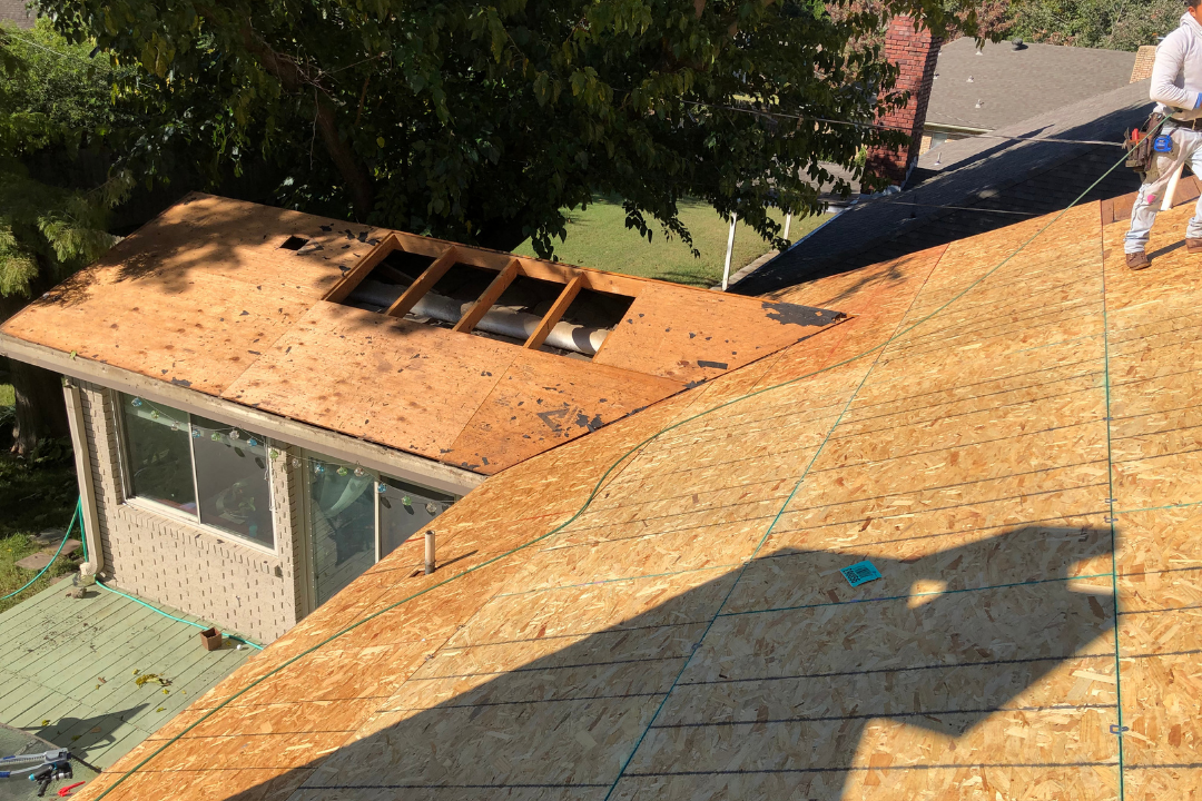 Full new OSB decking installed — snap lines visible, crew member on roof, old lower section with skylight cutouts, sunny Tulsa summer