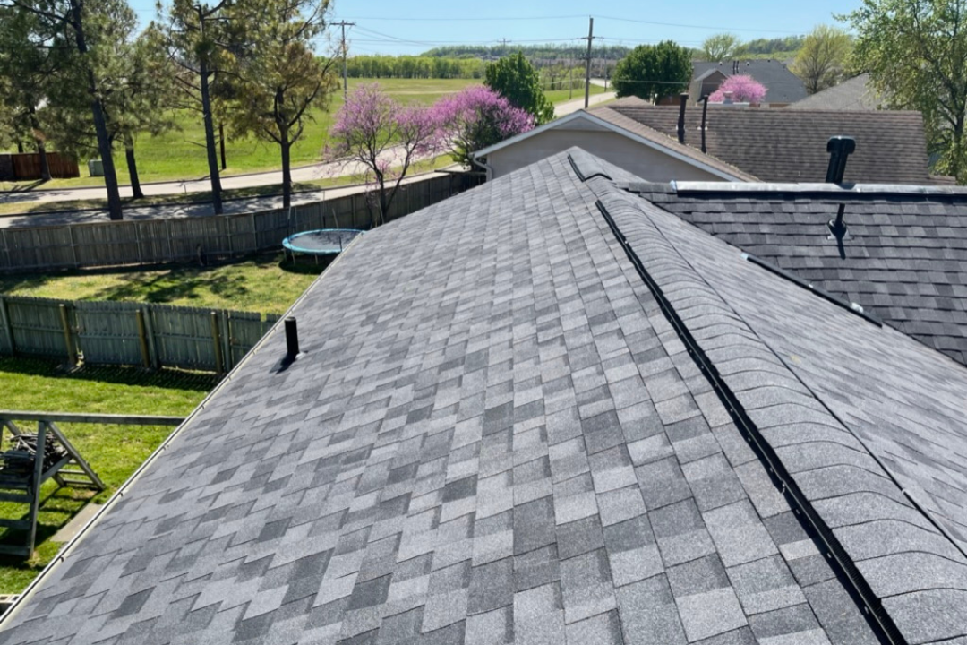 Clean finished gray roof with full ridge vent — pipe jacks, perfect install, redbud trees blooming, spring Oklahoma sky