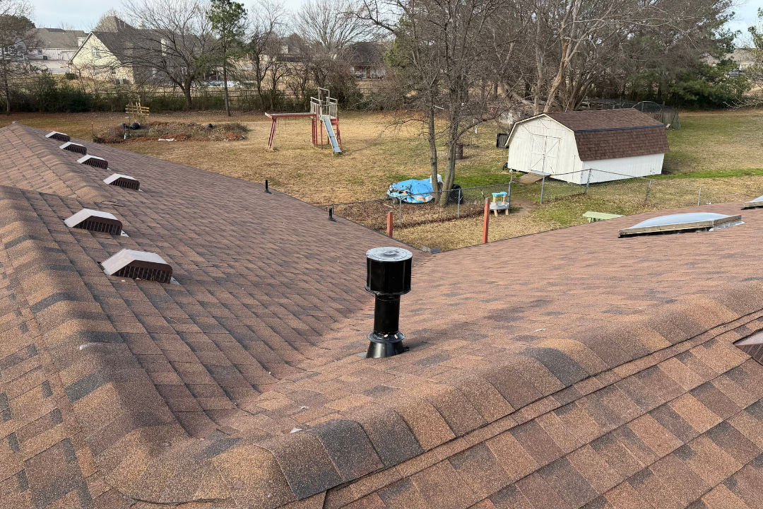 Clean completed brown shingle roof — row of box vents, black pipe jack, skylights, Tulsa suburban backyard with playset