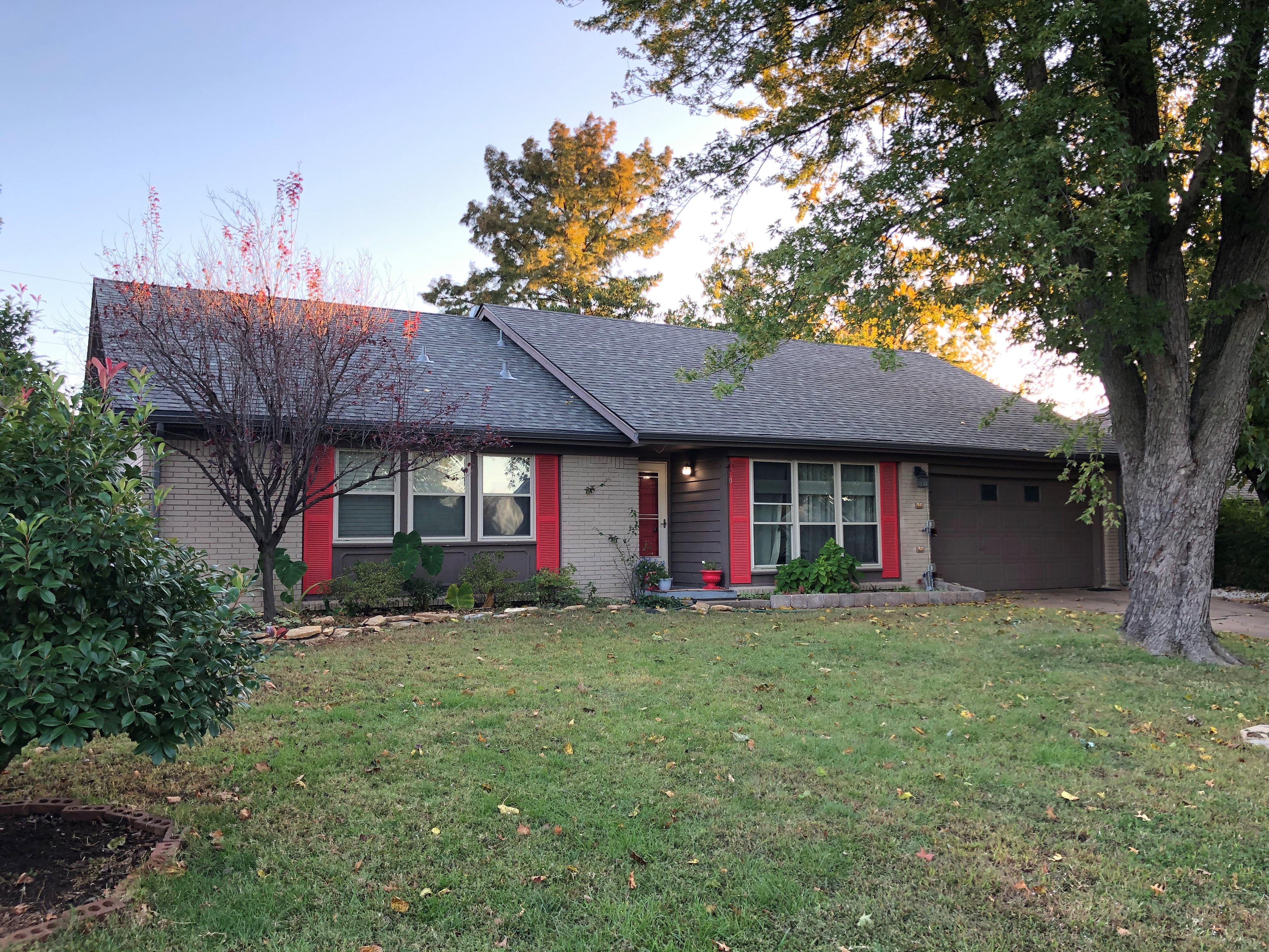 Completed dark shingle roof on Tulsa brick home — red shutters, mature trees, fall colors, golden hour. Happy ending after claim paid.