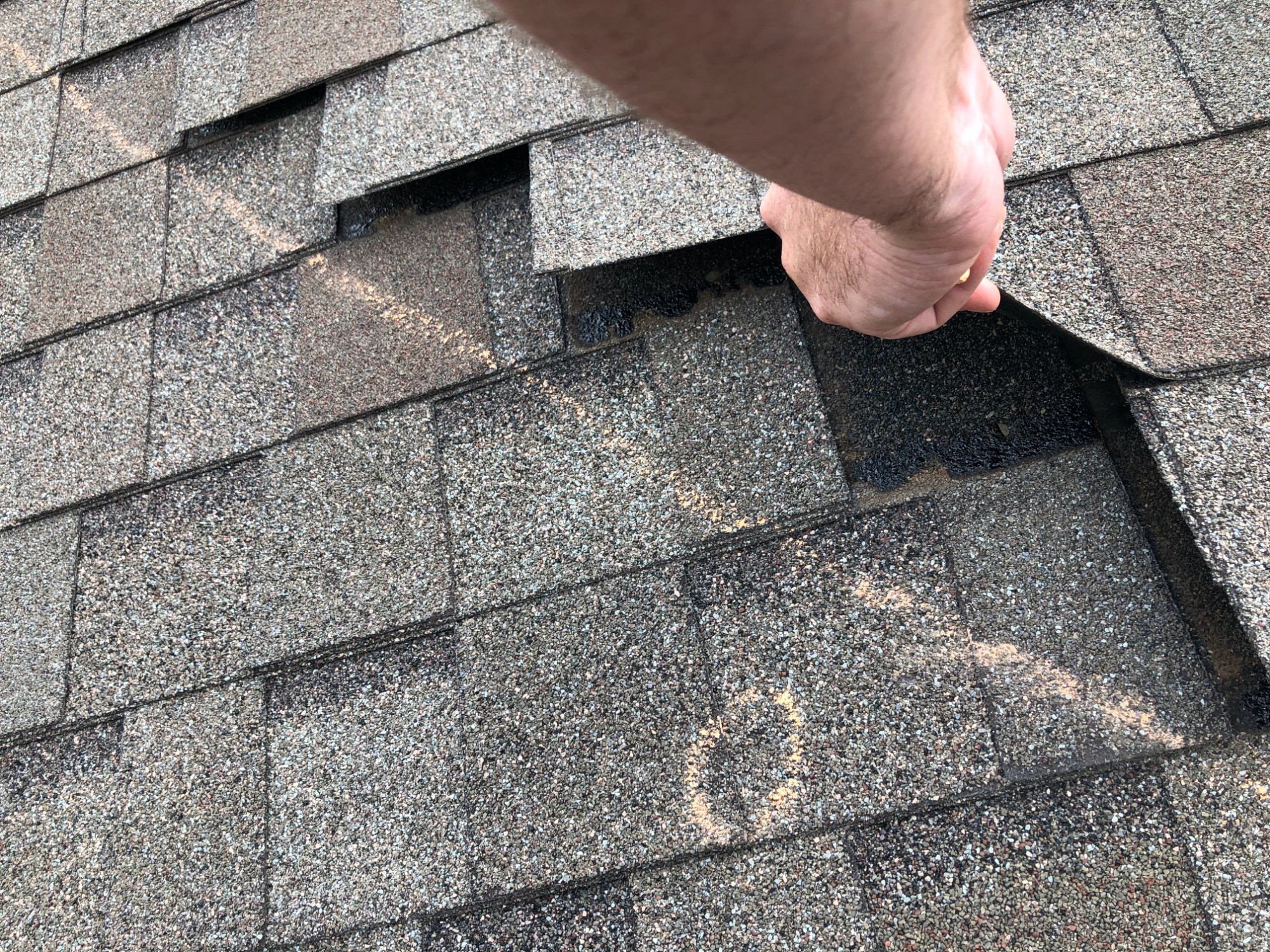 Rob's hand lifting damaged shingle — wedding ring visible, chalk marks on surrounding shingles